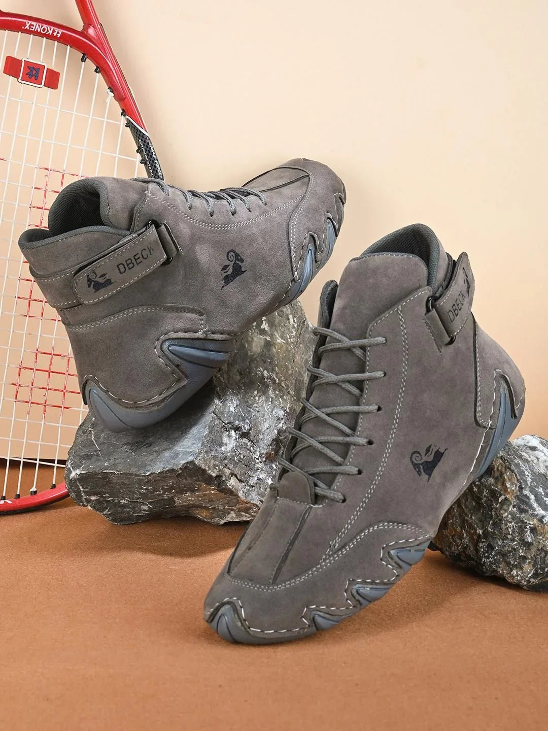Gray suede high-top sneakers with unique stitching on rocks, red tennis racket in background